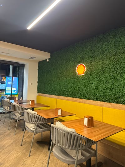 The image depicts the interior of a modern cafe, possibly a rest stop, with a Shell logo prominently displayed. The cafe features a long, communal table with a light wood finish, illuminated by a bright overhead strip light. Yellow banquette seating runs along one wall, contrasting with the natural wood tones. Several grey chairs with woven seats and backs are arranged around the table. A faux green hedge wall covers a portion of the ceiling and one wall, adding an element of natural decor. A neon Shell logo, shaped like a scallop shell, glows on the green wall, indicating the brand affiliation. Small wooden dispensers, likely for napkins, are mounted on the table. In the background, a large screen displays colorful graphics and text, possibly advertising or menu information. A security camera is visible mounted on the wall near the green hedge. The overall ambiance suggests a clean, contemporary, and functional space. No people are clearly visible, and the time of day is indeterminate due to interior lighting.