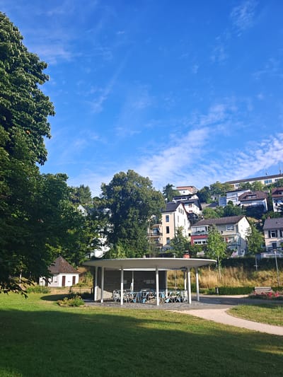 The image depicts a sunny day in Bad Schwalbach, Germany, with a clear blue sky dotted with wispy white clouds. In the foreground, a well-maintained green lawn leads to a modern open-air shelter with a white roof supported by pillars. The shelter has a dark brick wall in the back and appears to house several bicycles. To the left of the shelter, a small, white, gabled building with dark brown doors and windows is nestled amongst trees. Behind the shelter, a gravel path winds through the landscape, and a park bench with a small flower bed is visible on the right. The background features a steep hillside covered with dense green trees and a cluster of residential buildings with varying architectural styles and colors. The overall scene suggests a peaceful, residential area within the town.