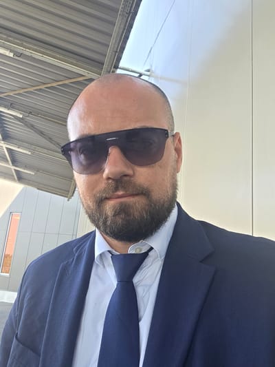 A bald man with a beard and sunglasses is wearing a dark blue suit jacket over a light blue dress shirt and dark blue tie. He is outdoors with a corrugated metal roof structure overhead and a pale, textured wall to his right. The background also shows a section of a light-colored building with a small, orange-tinted window. The man is looking towards the camera.