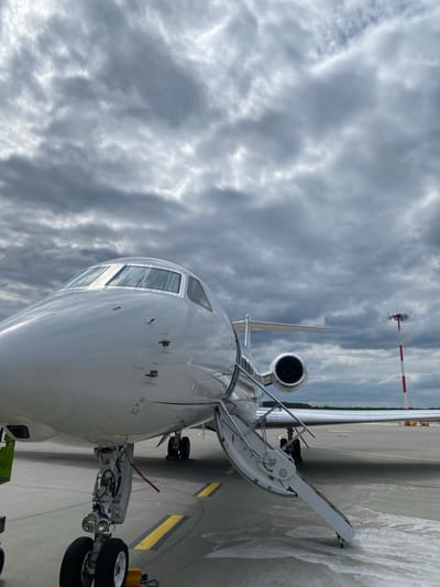 The image displays a close-up, low-angle view of a white private jet parked on an airport tarmac in Skulte, Latvia. The aircraft is seen from its front-right side, with its nose pointed slightly away from the viewer, set against a heavily clouded sky.

The primary subject is a sleek, white business aircraft. Its cockpit windows are prominently visible on the nose, reflecting the grey sky. Below the cockpit, the front landing gear with its wheels is firmly on the tarmac. On the right side of the fuselage, the main cabin door is open, with its integrated stairs extended and lowered to the ground, indicating readiness for boarding or disembarkation. Towards the rear of the aircraft, a portion of an engine intake is visible on the right side, along with the leading edge of a wing and a part of the vertical stabilizer (tail fin).

The setting is an airport tarmac, characterized by the grey paved surface and clear yellow painted lines. In the background, a tall red and white striped pole, possibly a meteorological mast with an anemometer, stands against the horizon. A faint line of dark green trees suggests the airport's perimeter.

The weather appears overcast or heavily cloudy during daytime, with various shades of grey clouds dominating the sky. No individuals are visible in the scene. Several warning labels are visible on the aircraft's fuselage near the cockpit windows in red text, including "DO NOT PUSH", "DO NOT TOUCH", "NO STEP", and "PITOT" near a small probe.