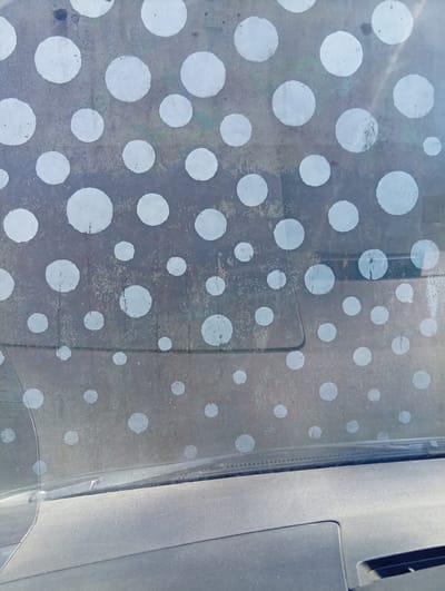 The image shows a close-up view of a car's windshield, which is covered in a pattern of white circular dots. The windshield appears to be dusty or grimy, with some areas of discoloration and streaking behind the dots. The dots themselves vary in size and are not uniformly spaced. The view is from inside the car, looking out through the windshield, with the dashboard and part of the interior visible at the bottom of the frame. The environment outside the car is not clearly discernible, but the lighting suggests daylight. The location is specified as Dirvonėnai, Lithuania.
