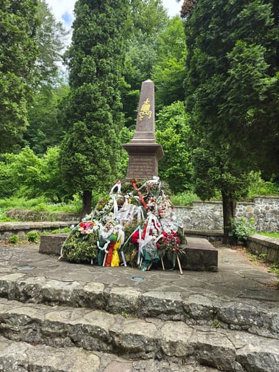 An outdoor, eye-level forensic photograph captures a stone monument situated amidst dense green foliage in Ribaritsa, Bulgaria, likely the Georgi Benkovski Monument. The monument consists of a dark, roughly square-based plinth supporting a tapering, obelisk-like stone shaft. A golden, stylized emblem, possibly depicting a religious or historical symbol resembling a bishop's mitre and staff, is prominently displayed near the top of the shaft. The plinth features an inscription in Cyrillic script, partially legible as "ГЕОРГИ БЕНКОВСКИ" and references to "1876" and "КОСТИЯ."

The base of the monument is heavily adorned with numerous memorial wreaths and floral arrangements. These tributes comprise a variety of fresh flowers, including red, white, pink, and yellow blossoms, interwoven with green foliage. Several wreaths are affixed with white ribbons, some bearing Cyrillic text, while others feature ribbons in the white, green, and red colors of the Bulgarian flag, along with yellow and orange accents. Some wreaths are supported by small wooden easels.

The monument is set on a paved area composed of irregular, light-colored stone slabs, which also form a path. In the immediate foreground, three weathered, rough-hewn stone steps, light grey with patches of lichen, lead up to the monument's platform. The area is enveloped by lush vegetation; tall, slender evergreen trees (resembling cypresses) stand to the right and left of the monument, while a dense canopy of various deciduous and evergreen trees forms a verdant backdrop. A low, dry-stacked stone wall is visible in the mid-ground to the right. The scene is illuminated by natural, diffused daylight, casting no strong shadows. No individuals are present in the image.