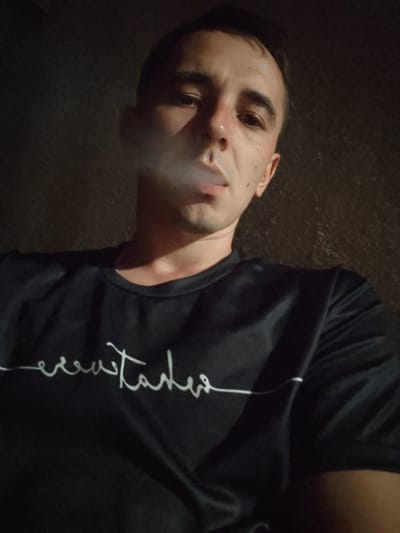A close-up, low-angle selfie captures a man exhaling smoke. He is wearing a black t-shirt with white script lettering across the chest, partially obscured by the smoke. The man's face is illuminated from the front, with the background a dark, textured surface, suggesting an indoor or dimly lit outdoor setting. The image is slightly grainy, and the focus is primarily on the man's face and upper body. The overall impression is informal and candid. The image is located in the city of Shkurupiivka, Ukraine.