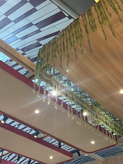 The image shows an interior view of a ceiling with decorative elements. A light-colored fabric canopy is stretched between red beams, with recessed ceiling lights illuminating the space. Above this, a slanted wooden panel features artificial green vines hanging down. In the background, a portion of the ceiling displays a checkered pattern of light and dark rectangular tiles. The scene is likely a commercial or public space. The image does not depict any people or specific actions.