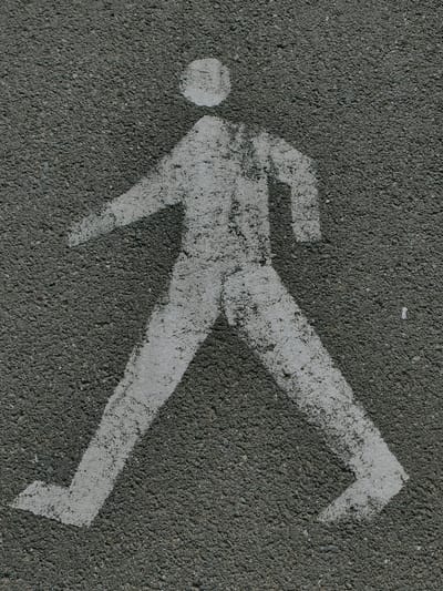 This is an eye-level, medium shot of a white pedestrian crossing symbol painted on dark asphalt. The symbol depicts a stylized human figure walking, with its left leg extended forward and its right leg slightly behind. The figure is composed of simplified geometric shapes: a circle for the head, a rectangular torso, and two lines for arms. The paint is somewhat worn and faded, revealing the texture of the asphalt beneath. The background is entirely the dark, granular surface of the asphalt, with no other objects or environmental features visible. This symbol is likely located on a sidewalk or roadway in the city of Murska Sobota, Slovenia, indicating a designated area for pedestrians to cross.