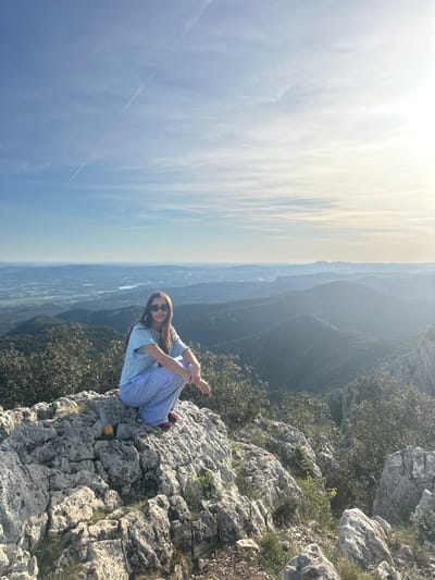 A medium shot captures a person sitting on a rocky outcrop. The person wears a light blue t-shirt, light blue pants, sunglasses, and red shoes. They sit cross-legged with arms resting on their knees, appearing relaxed. The rocky outcrop is covered with sparse vegetation and overlooks a vast landscape consisting of hazy mountains, valleys, and a distant city or town. The sky transitions from bright white near the horizon to a lighter blue higher up. The scene is located at a high vantage point above Bonnieux, France, given the expansive view. A small object, possibly a container, sits near the person's feet on the rocks. The image is rotated counter-clockwise approximately 90 degrees.