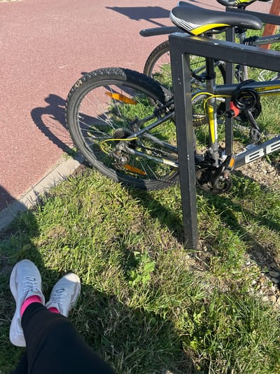 An outdoor scene shows two bicycles parked at a bike rack in Bałdowo, Poland. The bicycles are silver with yellow accents. One bicycle has a black seat with yellow stripes. Both bicycles are secured with a red and black cable lock. The ground is covered in green grass and a red path runs along the left side of the image. In the foreground, a person's legs are visible, wearing white sneakers and pink socks. The person appears to be sitting or resting. The sun is casting shadows across the scene.