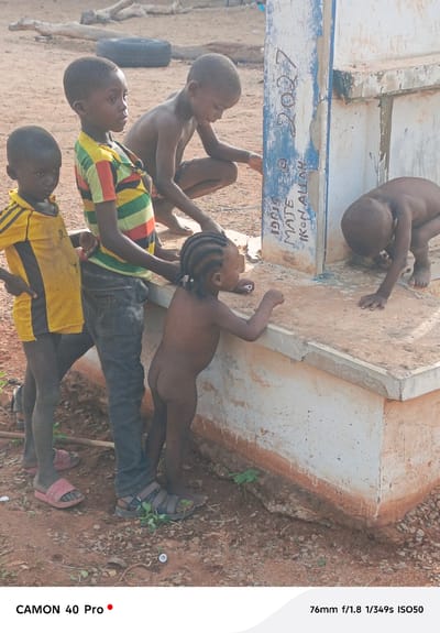 A ground-level shot depicts four young children gathered around a low, rectangular concrete structure. The setting appears to be in the city of Gidan Kogi, Nigeria, based on the signage on the concrete structure.

A boy in a yellow and black shirt stands on the left, next to a boy wearing a striped yellow, red, and green shirt and blue jeans. A small, nude girl stands in front of the boy in jeans, facing the concrete structure. A boy in the back has crouched down on the concrete structure as well. On the right side of the structure, another child is kneeling, and partially nude.

The concrete structure has some text in black ink. The text says: "IDRIS MATE IKON ALLAH" and "2007" is written above it. The ground is dirt.