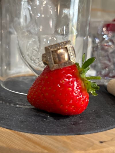 The image shows a close-up, angled shot of a single strawberry resting on a dark, textured surface, possibly slate, which is next to a wooden surface with vertical grain. The strawberry is vibrant red with visible seeds and a green stem with leaves. A ring, encrusted with small, sparkling stones and featuring a larger square centerpiece, is placed around the base of the strawberry, partially obscuring it. The background is blurred, revealing a clear glass object, possibly a jar or bottle, with light reflecting off its surface, creating bokeh effects. Hints of red objects are visible in the further background, also blurred. The lighting appears bright, with strong highlights on the strawberry and the ring, suggesting an indoor setting, possibly a kitchen or dining area. No text is visible in the image. The overall mood is still and somewhat luxurious, given the presence of the jeweled ring. The setting is not explicitly detailed, but the wooden element could suggest a rustic or natural aesthetic alongside the polished ring and fresh fruit.
