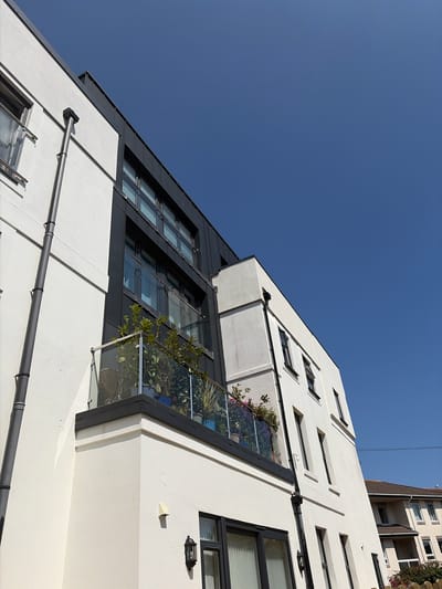 The image shows the exterior of a modern, white-rendered apartment building on a clear, sunny day. The building features dark-framed windows and a dark, angular roofline. There is a balcony with a glass balustrade, adorned with potted plants, extending from the upper floor. A dark drainpipe runs vertically down the side of the building. In the background, another residential building with a tiled roof is partially visible. The scene is set in Torquay, United Kingdom.