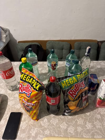 The scene, located in the city of Djigurovo, Bulgaria, displays a collection of consumable items arranged on a table covered with a light-colored tablecloth featuring a repeating leaf pattern. Objects present include: one partially full large plastic Coca-Cola bottle, one full 1.5-liter plastic Coca-Cola bottle, three partially full green plastic DEVİN branded water bottles, one partially full small green plastic bottle with a yellow cap, and two empty clear glass bottles consistent with alcohol containers, one retaining a blue label remnant and the other a red-gold cap. Two "MEGAPACK Ruffles" chip bags, identified as "Barbecue" and "Ketchup" flavors, are positioned on the table, both appearing largely full. To the right, a blue Red Bull multipack packaging containing at least two cans, and a white carton depicting strawberries are visible. A black smartphone is placed face down on the tablecloth on the left. The background features a plain white wall above a light-colored sofa or bench fitted with dark green tufted cushions. A dark backpack rests on one of the sofa cushions. No individuals are visible within the frame.