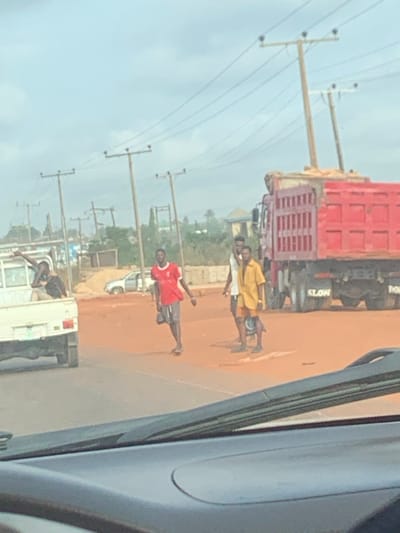The image captures an outdoor street scene in Issele Uku, Nigeria, under an overcast or hazy sky. The perspective is from inside a vehicle, with its dashboard and windshield wipers visible in the lower foreground.
The ground surface transitions from a dark grey paved section in the lower-left to a dominant expanse of red-brown dirt or sand across the mid-ground, featuring some darker, possibly wet, patches.
On the left, a white utility truck with an open bed is partially visible. A dark-skinned male, wearing a dark top, is seated in the truck bed with his right arm raised and extended to the left. Behind this truck, a silver sedan is partially visible on the dirt shoulder.
To the right of the white truck, three dark-skinned males are present on the dirt road. The leftmost male wears a red short-sleeved shirt with white trim, dark shorts, and dark shoes, walking towards the left of the frame while holding a dark object in his left hand. To his right, two other males stand or walk; one wears a light-colored short-sleeved shirt and dark shorts, and the other, positioned slightly behind him, wears a yellow short-sleeved shirt and dark shorts, also holding a dark object in his left hand.
On the right side of the frame, a large red dump truck, heavily laden with a light-colored granular material, occupies the roadway. The word "SLOW" is visible on its rear mud flap.
The background features multiple wooden utility poles supporting numerous overhead power lines that traverse the scene horizontally. Distant buildings and sparse vegetation are visible on the horizon beneath the light grey sky.