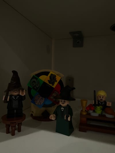 The image displays a static arrangement of Lego Harry Potter minifigures and associated accessories on a white surface, consistent with a shelf or display area, located indoors in McAllen, United States of America.
On the left, a Harry Potter minifigure, identifiable by his glasses, black uniform, and dark brown sorting hat, stands on a small, round, three-legged brown Lego stool. He holds a dark brown wand in his right hand.
In the center foreground, a Professor Minerva McGonagall minifigure, characterized by her green robe, black witch hat, and spectacles, stands holding a black wand in her right hand. Positioned behind this minifigure is a large, light brown circular Lego structure, featuring a black central disc surrounded by four colored square panels depicting the Hogwarts house crests: a green panel with a coiled snake (Slytherin), a yellow panel with a lion (Gryffindor), a blue panel with an eagle (Ravenclaw), and a red panel with a badger (Hufflepuff).
On the right, a Draco Malfoy minifigure, distinguished by his blonde hair and black uniform, stands behind a dark brown Lego table. The table supports a golden goblet, a stack of green cylindrical pieces, and two red spherical pieces. The Malfoy minifigure holds a black wand in his right hand.
The background consists of plain white walls. In the upper right corner of the background, a dark L-shaped metal bracket, secured by two visible screws, is affixed to the wall. The scene is dimly lit.