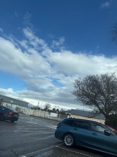 This image depicts an outdoor parking area in Oberhausbergen, France, under a partly cloudy sky with visible patches of blue. The foreground on the right shows the rear passenger side of a dark teal BMW 3 Series Touring, parked diagonally across a white-striped asphalt parking space. Behind it, a portion of a red vehicle is visible. To the left, a dark grey Tesla Model S (license plate EC-388-NQ) is parked facing away from the viewer on a wet asphalt surface. A large, bare deciduous tree with multiple branches occupies the mid-right. In the background, a modern grey building with a flat roof and dark upper section is visible behind a white security fence. Several light poles and a white van are also present behind the fence. Further in the distance, construction cranes are visible against the horizon line. No individuals are present in the scene.