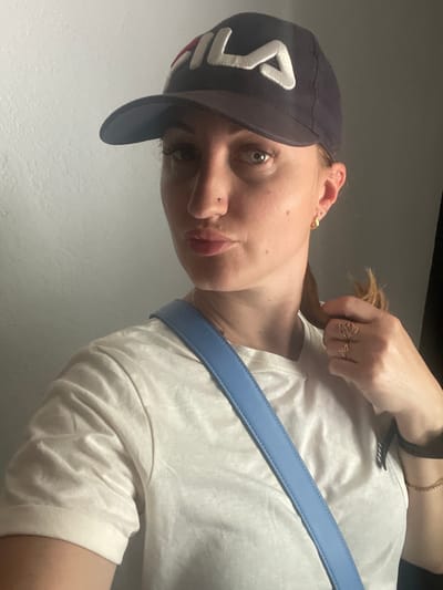 The image features a young woman, likely in her late teens or early twenties, with fair skin and light-colored eyes. She is wearing a dark baseball cap with a white logo that appears to say "FILA." She is also wearing a light-colored t-shirt and a light blue strap, possibly from a bag, is visible across her chest. She has a piercing on her nose and is wearing rings on her fingers. Her hair is pulled back. The lighting suggests it could be daytime, though the specific time is unclear. The background is a plain, light-colored wall, indicating an indoor setting. The overall mood is casual and relaxed. There is no discernible weather or location cues beyond the simple indoor environment. The image appears to be a selfie, given the angle and focus on the subject.