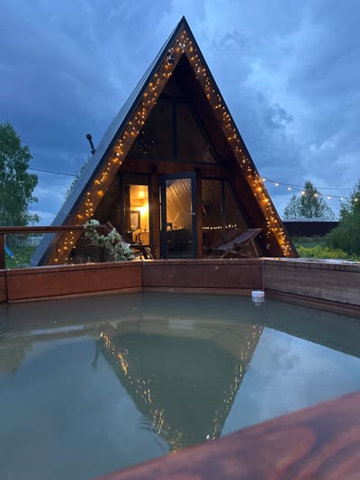 The image depicts a modern A-frame cabin in Малеевка, Russia, at dusk. The cabin is situated next to a body of water, likely a pond or lake, with a portion of the cabin appearing to be built directly adjacent to it. The exterior is adorned with string lights that illuminate the roofline and reflect in the water. The cabin features large glass windows and doors. Interior lighting is visible through the glass. There is a wooden deck or platform extending from the cabin's structure towards the water. The sky is overcast with dark blue and gray clouds. Trees are visible in the background.