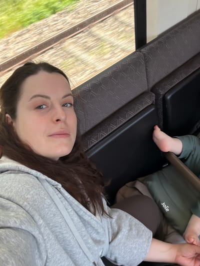A woman lies on a train seat next to a baby in a Joie-branded highchair. The train window shows a blurred view of the outside, potentially near Odernheim am Glan, Germany. The woman has brown hair, blue eyes, and is wearing a grey hoodie. The baby's foot is visible. The train seat is patterned and dark.