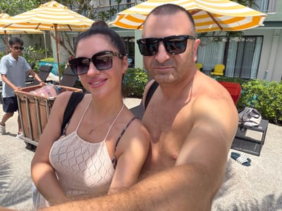 A man and a woman, wearing sunglasses and casual attire, pose for a selfie outdoors. The man is shirtless, while the woman wears a white knitted top and a necklace. Behind them, a young boy in a blue shirt and dark shorts pushes a cart. Yellow and white striped umbrellas are visible, along with lounge chairs and a swimming pool. The setting appears to be a resort or hotel in Bang Muang, Thailand, with lush greenery and buildings in the background. The ground is paved with light-colored pebbles.