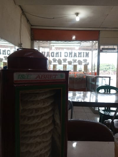 The indoor shot reveals a dining area, likely a restaurant, featuring tables and chairs. A tall, maroon dispenser with the label "I & E ABRET" and "PERUSAHAAN KERURUM IKAN" houses neatly stacked, rope-like snacks. Behind, a window displays "MINANG INDAH MINANG," suggesting the establishment's name and possibly its regional cuisine. Rows of bowls are visible on shelves within the restaurant. A light fixture illuminates the scene. The overall impression is of a simple, possibly family-run, eatery.