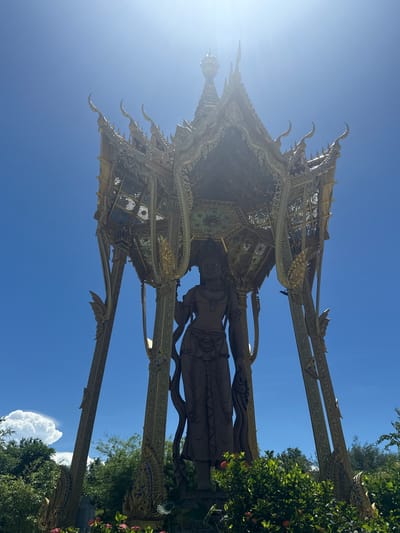 The image depicts a golden statue of a figure, likely a deity, standing beneath an ornate, open-air pavilion. The statue appears to be made of a dark, reflective metal. The pavilion has a multi-tiered roof with upward-curving eaves and elaborate carvings. The surrounding environment includes green trees and shrubs. The sky is bright blue, suggesting a sunny day. The location is Bang Pu Subdistrict Municipality, Thailand.
