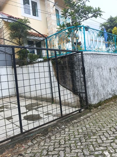 The image depicts an exterior view of a residential property and its immediate surroundings in the city of Pô Kô, Vietnam. The scene is observed from a slightly low angle, looking upwards towards a multi-level building.

In the foreground, a dark grey, interlocking paver pathway or roadway is visible, featuring notable moss growth within the interstices. A black metal grid-patterned sliding gate, constructed from square tubing, is partially open, with its right panel angled towards the viewer's right. Behind the gate, a concrete slab driveway exhibits several irregular dark wet patches.

To the left of the driveway, a white-painted retaining wall with vertical dark streaks and a faint grid pattern supports elevated ground. To the right, a rough-textured grey retaining wall, also showing dark staining and moss at its base, separates the property from the foreground pathway.

The main subject is a multi-story building with light yellow/cream painted walls. The upper level features a prominent white-framed window with a decorative grid, and an adjacent section of wall is clad in light-colored, stacked stone-like tiles. Above a lower, partially obscured window, a brown-tiled awning is visible. An elevated terrace or balcony is present, featuring a distinct turquoise blue railing adorned with gold-colored oval decorative accents in a repeating pattern.

Vegetation includes a small leafy bush behind the left retaining wall and a larger tree with broad leaves positioned on the elevated terrace, partially obscuring the turquoise railing. Additional dark green foliage is visible further in the background against what appears to be an overcast sky. Utility wires are present across the upper section of the image, spanning in front of the building. No individuals or animals are present in the frame.