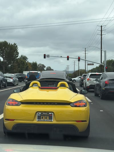 This is a street-level, eye-level, full-shot photograph taken from inside a vehicle, looking out at traffic on a highway. The image captures a yellow Porsche convertible in the foreground, with its rear facing the camera. The license plate reads "CAIA" and indicates "McKenna" and "California" with an expiration of "JUN". The car has its brake lights illuminated. In the background, there is a multi-lane highway with moderate traffic, including other cars, SUVs, and a Tesla. Traffic lights are visible, with some displaying red signals. A street sign indicating "Irvine Blvd" is also present. The sky is overcast and gray, suggesting a cloudy day. The environment appears to be urban or suburban, characterized by roads, traffic signals, and utility poles with power lines. The location is identified as Irvine, United States of America.
