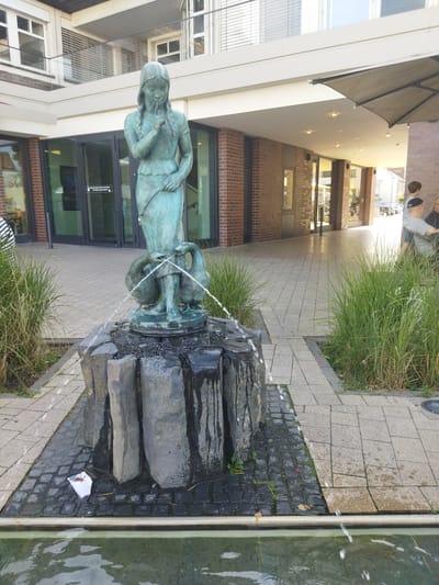 The image shows a bronze statue of a young girl with pigtails, her finger held to her lips in a shushing gesture. She is depicted standing with two geese, one on each side, with water spraying from their beaks. The statue is situated on a rough-hewn stone fountain base, which is surrounded by water. Tall green grasses are planted around the fountain. The fountain is located on a paved area with a tiled walkway leading away from it, and a building with large glass doors and brick accents is visible in the background. The overall scene suggests an outdoor public space in a city. The location is identified as Monheim am Rhein, Germany.