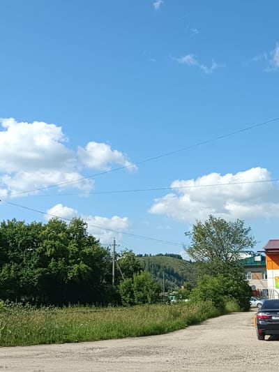 The image shows a daytime scene in a rural or suburban area with a blue sky and scattered white clouds. In the foreground, there is a dirt road curving to the right. To the left of the road, there is a grassy field with some wildflowers. Behind the field, there are dense green trees and a utility pole. Further in the background, there are hills covered with trees and some buildings with colorful roofs. A black car with a visible license plate is parked on the right side of the road. The overall impression is a bright, sunny day in a somewhat undeveloped area. There are no people visible in the image.