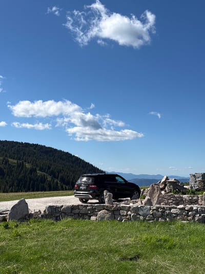 A black SUV is parked on a gravel road at the edge of a grassy hill. The car is facing right, with its rear end visible. In the foreground, a low stone wall made of rough-hewn rocks runs across the frame. Beyond the wall, the grassy hillside slopes down. In the background, a dense forest of evergreen trees covers a mountain. The sky above is a clear blue with scattered white clouds. The scene appears to be a natural landscape, possibly in a mountainous or hilly region. The license plate of the car reads "PB 1234 ME".