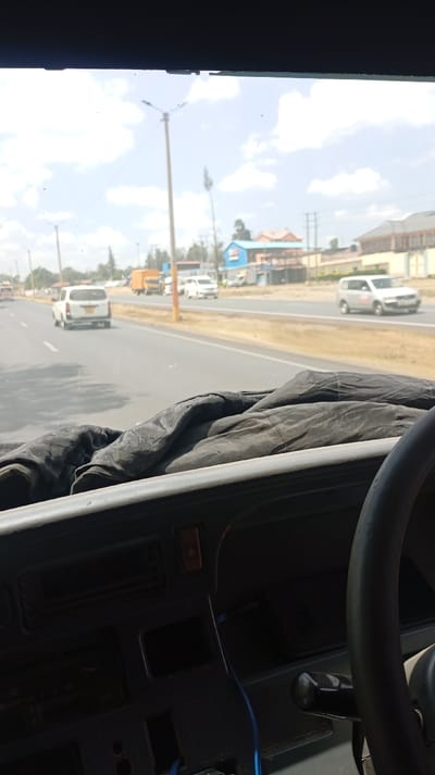 The image depicts a view from inside a vehicle, focusing on the dashboard and steering wheel. Outside the windshield, a road stretches into the distance with several vehicles moving along it. Buildings and utility poles line the roadside under a slightly cloudy sky. The interior of the vehicle is dimly lit, contrasting with the bright daylight outside. A dark cloth covers the dashboard.