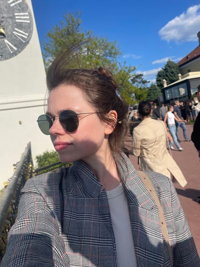 The original image, presented horizontally, shows a young woman in the foreground against a blurred backdrop of an outdoor public space in Petrovaradin, Serbia. When viewed upright, the scene depicts a clear blue sky with scattered white clouds, indicating bright daylight.
The primary subject, a woman with dark brown hair pulled back, is visible from the chest up, facing to her left in profile. She wears dark-rimmed sunglasses and a stylish grey plaid blazer with subtle red and black patterns, layered over a white ribbed top. A light beige belt is visible beneath her blazer. Her expression, partially obscured by the sunglasses, appears calm and relaxed.
The background, rendered out of focus, reveals several people in various activities, suggesting a lively public area. One person in a light beige coat is seen directly behind the main subject. Further back, other figures are walking or standing on a reddish-brown paved surface, surrounded by green trees and parts of buildings, possibly shops or cafes.
Distinctive details in the foreground include a large, grey clock face with Roman numerals (such as I, II, V, X) and gold hands, partially visible on the left side of the frame. Below the clock and extending to the right, a railing is covered with numerous love locks, predominantly gold and silver, some with red accents. These specific elements, combined with the location context, point to the famous clock tower area of the Petrovaradin Fortress. The bright sunlight casts strong shadows, reinforcing the impression of a sunny day. No legible text is visible on any signs or objects in the background.