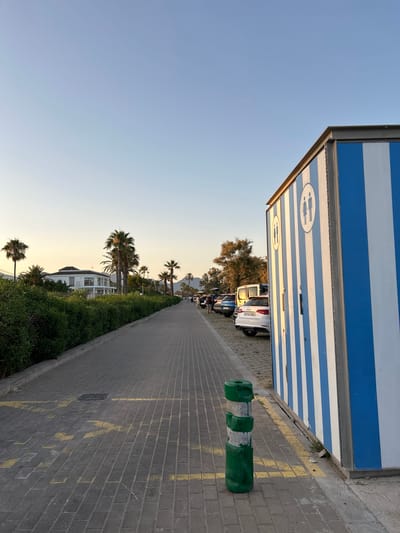 The image shows a paved walkway next to a building with blue and white striped siding. There are two circular symbols on the building, each depicting a man and a woman, indicating a restroom facility. A line of parked cars is visible behind the building, including a white SUV. To the left of the walkway, there is a strip of green vegetation, with several palm trees and a white building with a tiled roof. The sun is low in the sky, casting long shadows and creating a warm, golden light, suggesting it is either early morning or late afternoon. The sky is clear and blue. A green and white bollard stands on the walkway. Yellow lines are painted on the brick paving in places. The scene appears to be in a coastal or tourist area, possibly a promenade or parking lot near a beach. There is no clear activity taking place other than the presence of parked vehicles. The overall mood is peaceful and serene, bathed in the soft light of sunset or sunrise. The license plate on the white SUV is partially visible, reading "9704 AML".