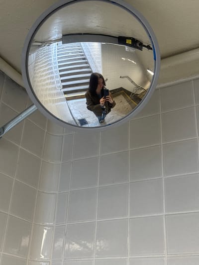 The image displays a large, circular convex mirror with a grey frame, mounted on a light-colored ceiling. The mirror reflects a single individual, appearing as a female with dark hair, wearing a dark olive-colored jacket over a lighter top, blue jeans, and light-colored footwear. A chain-link strap is visible over her right shoulder. She holds a dark-colored smartphone with both hands, positioned to capture her reflection in the mirror, with wired earbuds dangling from the device.
The background reflected in the mirror details a public stairwell within what appears to be a transit station. This area features a flight of light-colored concrete stairs with defined treads and risers, ascending towards the upper left from the reflected perspective. Metal handrails line the stairwell. The walls within the reflected area are clad in light-colored square tiles, matching the tiling in the immediate foreground. A small floor grate is visible near the base of the reflected stairs. Overhead in the reflection, a dark-colored piece of equipment or light fixture with a yellow label is mounted.
The immediate environment surrounding the mirror, from the perspective of the original photograph, consists of light grey or off-white square tiles with white grout, covering the walls and a curved corner. This specific architectural style, including the tiled passageways and stairwells, is characteristic of public transit infrastructure in the city. The location is consistent with a Port Authority Trans-Hudson (PATH) station in Hoboken, New Jersey, United States of America.