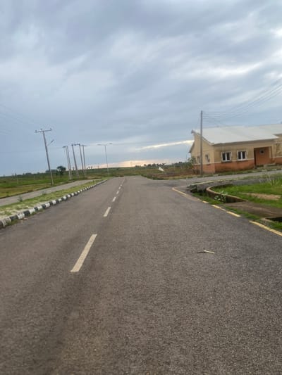 The media file depicts a street scene in Bulamare, Nigeria. The foreground is dominated by an asphalt road with white dashed lines marking lanes. To the left of the road, there is a curb with alternating black and white markings, followed by a strip of grass and then more of the road. In the distance, on the right side of the road, a modern residential building with beige and orange-colored walls and multiple windows is visible. Beyond the building, there are utility poles with overhead electrical wires stretching across the landscape. The sky is overcast with a mixture of gray and white clouds, suggesting a cloudy or possibly hazy day. The time of day appears to be late afternoon or early evening, given the subdued lighting. The scene is quiet and appears to be in a developing residential area, with the road suggesting accessibility and infrastructure. There is no visible text in the image.