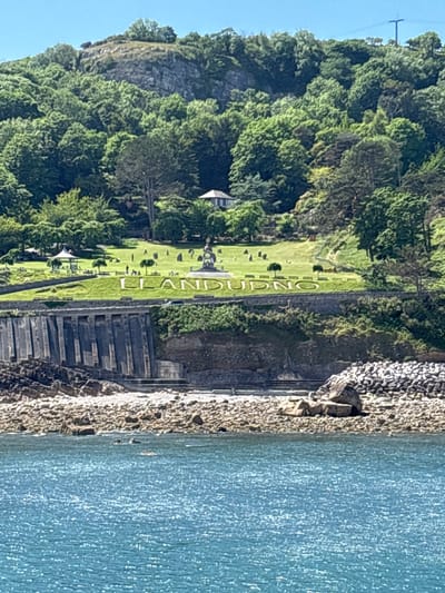 This is a landscape photograph taken in Llandudno, United Kingdom, featuring a hillside prominently displaying the town's name in large white letters on a grassy slope. The hillside is covered with dense green trees and vegetation. At the base of the slope is a stone wall with a staircase leading down to the water's edge. The water is a light blue color and appears to be part of the sea or ocean. The photograph is taken from an elevated position, looking down at the scene.