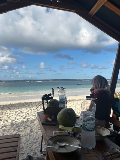 In the city of A R A, Indonesia, an outdoor dining area on a sandy beach overlooks the ocean. A person wearing a headscarf is seated at a wooden table, facing away from the camera and towards the water. On the table are two green coconuts, a partially opened bottle of Crystal water, a bowl with a spoon and fork, and a pair of glasses. The ocean is a vibrant turquoise, with small waves lapping at the white sandy shore. In the distance, several people are visible walking along the beach and a small boat is moored in the water. The sky is partly cloudy, with dramatic formations of white and grey clouds against a bright blue backdrop. The scene is framed by the wooden structure of a shelter, suggesting a relaxed and picturesque setting.