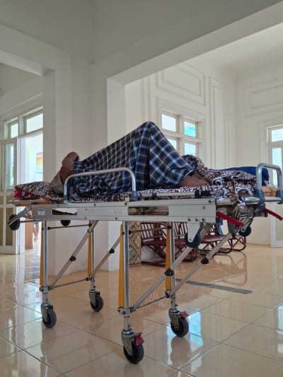A person is on a medical stretcher in what appears to be a hospital or clinic room. The person is partially covered with a blue and white checkered blanket and a patterned cloth. The stretcher is silver with yellow supports and black wheels. The room has light-colored tile flooring, white walls, and windows allowing natural light. In the background are some chairs. The location is Uleereuleung, Indonesia.