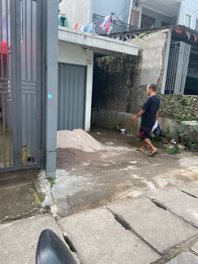 A man wearing a black t-shirt, red shorts, and black sandals walks to the left in front of a building. He is carrying a plastic bag. The building is a two-story residence, with a grey facade and a corrugated metal garage door. The image was taken in Bojongsoang, Indonesia, at the coordinates -6.979, 107.635. There is a pile of dirt on the ground in front of the garage door. There is a green plant growing in the dirt. The man is walking on a concrete surface. There are a series of drainage grates to the right of the man. There is a small blue container to the right of the garage door. The scene appears to be a residential neighborhood.
