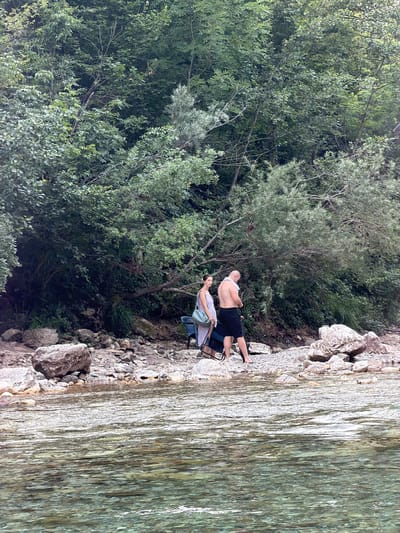 Two people are seen from behind, sitting on chairs by a rocky riverbank. The person on the left, a male, is shirtless and wearing black shorts, with a white towel draped over his shoulders. He is leaning back in his chair, looking towards the river. The person on the right, a female, is wearing a light-colored dress and is seated in a folding chair with a blue bag next to her. She appears to be looking down.
The scene is a lush, green forest on the left side of the image, bordering the rocky terrain. The river, on the right, has clear, shallow water with a visible riverbed. The sunlight reflects off the water's surface, creating a shimmering effect. The lighting suggests it is daytime, possibly mid-afternoon, and the weather appears calm. The location is identifiable as Spiljani, Bosnia and Herzegovina, based on the provided context. There is no visible text in the image. The overall mood seems relaxed and peaceful, with the individuals enjoying a moment by the water.