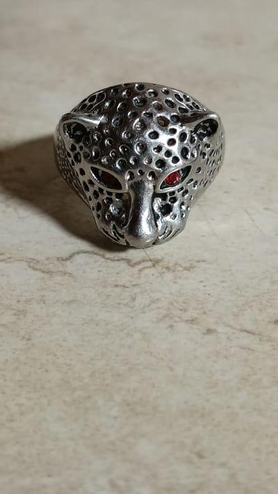 The image shows a close-up, high-angle shot of a silver ring shaped like a leopard's head, placed on a textured, light-colored surface, likely a countertop or table. The ring is detailed, with perforations resembling a leopard's spotted pattern covering its surface. Two small red stones are visible, one in each eye socket of the leopard's head, adding a vibrant contrast to the silver. The ring is positioned on the left side of the frame, with its face angled towards the right. The background is out of focus, highlighting the ring as the primary subject. There is no visible text, and the lighting suggests an indoor setting, possibly during the day. The overall mood is neutral, with the focus solely on the craftsmanship and appearance of the ring. The location context of Λεχαινά, Greece, is provided separately and does not appear to be directly indicated by visual cues within the image itself.