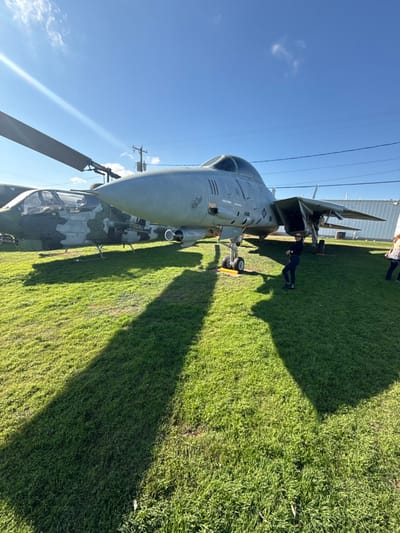 A close-up captures two military aircraft outdoors on a grassy area under a bright sky. One is a helicopter, featuring camouflage patterns, prominently displaying its rotor blades. Adjacent is a jet fighter, its grey fuselage detailed with visible cockpit, landing gear, and intricate wing structure. A silhouette of a person is observed around the jet, enhancing the aircraft's scale.