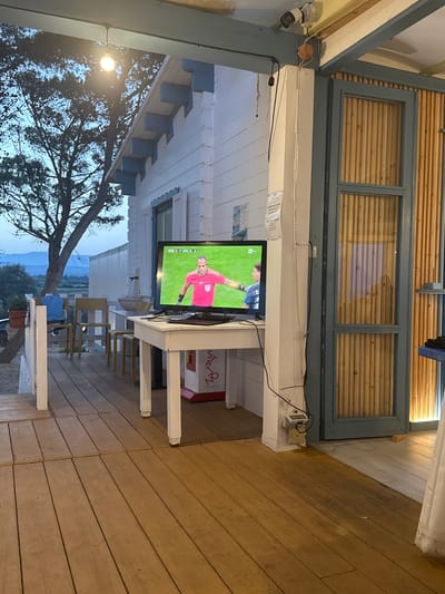 The image depicts a covered outdoor patio with a wooden floor and a white building with blue trim. A television is placed on a white table, showing a soccer match with a referee in a pink shirt and another player in a blue and black jersey. There are several chairs and a plant on the patio. In the background, there is a landscape with trees and hills under a twilight sky. The scene appears to be in Orosei, Italy, based on external information not present in the image itself.
