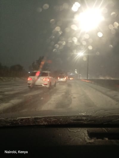 The image captures a nighttime or late evening scene from inside a vehicle, looking out through a rain-streaked windshield onto a multi-lane highway. Heavy rain is evident from the numerous water droplets on the glass, which cause a bokeh effect from distant lights, and the wet, reflective road surface.
In the midground, several vehicles are visible, driving in the same direction. A prominent white SUV, with its red taillights illuminated, is in the left lane. Further ahead, other cars and a van can be seen, their lights glowing faintly through the rain and atmospheric haze. Streetlights along the road cast bright, starburst-like flares, illuminating parts of the scene and contributing to the atmospheric glow. The road itself is dark and slick with water, reflecting the ambient lights and the vehicle's own path.
To the left of the road, dark shapes of trees or roadside foliage are discernible, while to the right, a barrier or median separates the lanes. The sky is dark and overcast, typical of a rainy night. The dashboard or lower part of the car's interior is visible at the bottom, showing more water residue and texture. The overall activity depicted is driving in challenging, rainy conditions. The visible text in the bottom left corner of the image states "Nairobi, Kenya."