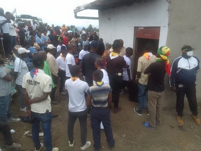 A large group of people is gathered outdoors next to a small, simple building. Many wear neckerchiefs suggesting a scout troop or similar organization. Some are entering the building through a doorway. The crowd is diverse, with individuals of varying ages and dress. A masked person in a tracksuit stands to the right, near the building. The setting seems somewhat rural, with a sloping terrain and trees visible in the distance.