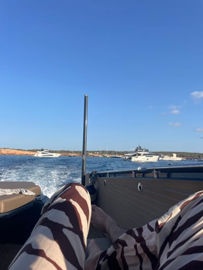 The image captures a first-person perspective from the stern of a boat in motion within the coastal waters of Formentera, Spain. The foreground displays the lower extremities of an individual, clad in trousers featuring prominent light and dark brown, irregular horizontal stripes. Bare feet are visible. To the left, a portion of a tan upholstered boat seat holds a folded light-colored textile item. To the right, the boat's side panel exhibits horizontal light brown slatting with circular metallic fittings. A dark grey cylindrical boat fender is partially visible.

The boat's movement is indicated by a white, foamy wake trailing in the blue water. A slender, dark grey metal pole extends vertically from the boat's floor, slightly left of the central axis. The midground reveals multiple vessels. A large white motor yacht is observed moving towards the far left. In the center-right, a multi-decked motor yacht, white with dark grey accents, is positioned in the water. Several other smaller sailing yachts and powerboats are visible at anchor or moving slowly in the distance. The background is defined by a reddish-brown rocky coastline adorned with green vegetation. The sky above is clear and bright blue, featuring scattered white cumulus clouds on the right. The scene is illuminated by natural daylight.