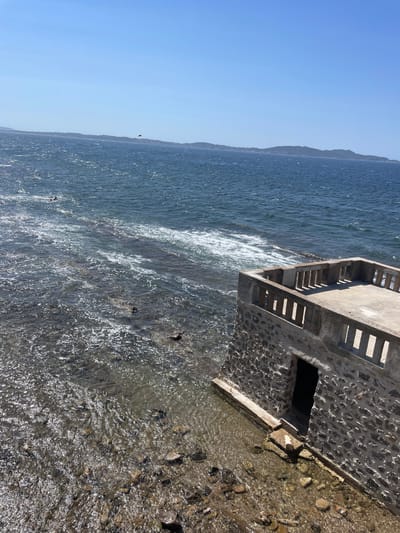 The image depicts a clear, sunny day with a bright blue sky over the Mediterranean Sea. In the foreground, a small stone building with a concrete balcony juts out over the water. The building has a rough stone facade and a doorway that appears to lead into darkness. The balcony has a concrete floor and a railing made of evenly spaced concrete posts. The water is choppy and appears shallow near the building, with rocks visible beneath the surface. Further out, the sea is a deeper blue, with distant hills or islands visible on the horizon. A small yellow object, possibly a parasail or kite, is visible in the far distance above the water. The scene is likely located in Carqueiranne, France, given the coastal setting and the architectural style of the building.