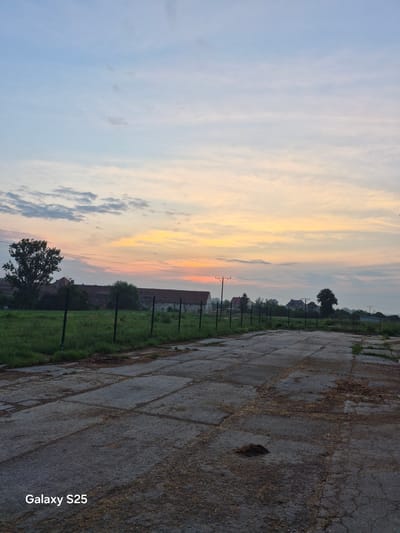 The image shows a concrete road leading into the distance with some debris scattered on its surface. To the left of the road is a grassy field enclosed by a metal fence. Beyond the field, there are several buildings, a large tree, and more vegetation. The sky above is filled with a gradient of colors, transitioning from pale blue at the top to shades of orange and yellow near the horizon, indicating either sunrise or sunset. The overall scene appears to be in a rural or semi-rural area. The "Galaxy S25" watermark is present in the bottom left corner.