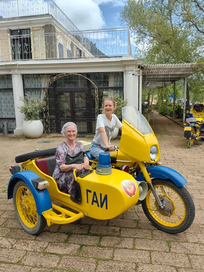 Two women pose with a vintage yellow and blue motorcycle with a sidecar on a paved area. An older woman sits in the sidecar with a dark bag. A younger woman sits on the motorcycle. The sidecar bears the Cyrillic letters "ГАИ." A building with columns and wrought iron details is in the background. The setting is identified as Luchistoe, Ukraine. Another similar motorcycle is partially visible in the background to the right. The lighting suggests a bright, overcast day.