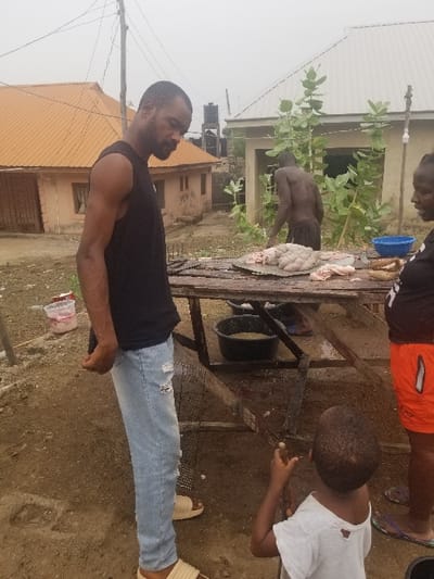 The scene takes place in Pati Ridge, Nigeria, at the coordinates 7.81 latitude and 6.704 longitude. A man wearing a black tank top and blue jeans stands facing a wooden table, his back to the camera. On the table is a large pile of raw, uncooked meat, presumably chicken,  and two blue plastic basins. A second man wearing a dark blue tank top and khaki shorts is standing by the table, facing away from the camera,  his back is to the viewer, he appears to be looking at the meat on the table. There is also a woman wearing a black shirt and orange shorts standing behind the table, facing away from the camera, her back is to the viewer, she is looking at the meat. A young boy wearing a white shirt and black shorts stands near the man in the black tank top, he is looking towards the meat on the table, holding a stick in his right hand. Behind the group of people is a brown wooden table and a large tree. There are several residential buildings in the background, with brown, orange, and white exteriors. The ground is dirt and a small red and white plastic container is visible in the foreground.  The scene is a street-side food market with a general sense of activity and business.  The weather appears to be overcast, with a faint light in the sky.
