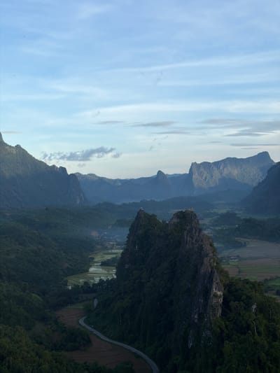 The image depicts a breathtaking panorama of mountainous terrain and a valley in the city of Naka, Laos. The foreground is dominated by a rugged, rocky peak covered in lush green vegetation. A winding asphalt road snakes its way through the valley below, bordered by agricultural fields, some of which appear to be rice paddies reflecting the sky. In the mid-ground, several other verdant, tree-covered mountains rise, creating a layered effect. The furthest mountains are shrouded in a soft, atmospheric haze, lending depth to the landscape. The sky above is a pale blue, streaked with wispy white clouds. Two small structures, possibly temples or observation posts, are visible atop the prominent rocky peak in the mid-ground. The overall scene evokes a sense of natural beauty and tranquility.